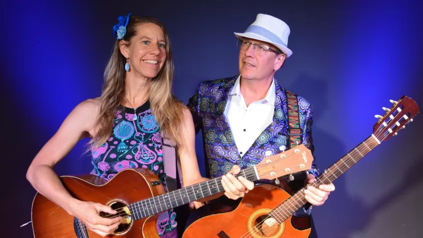 blue background with blonde woman and a man with hat on both smiling while holding a guitar