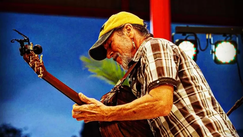 man in a baseball cap and brown striped short sleeve playing an acoustic guitar while standing in front of a night sky